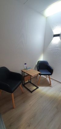 Wartebereich in der Podologie: Ein kleiner quadratischer Tisch mit je einem Stuhl rechts und Links und einer Wandleuchte im Hintergrund